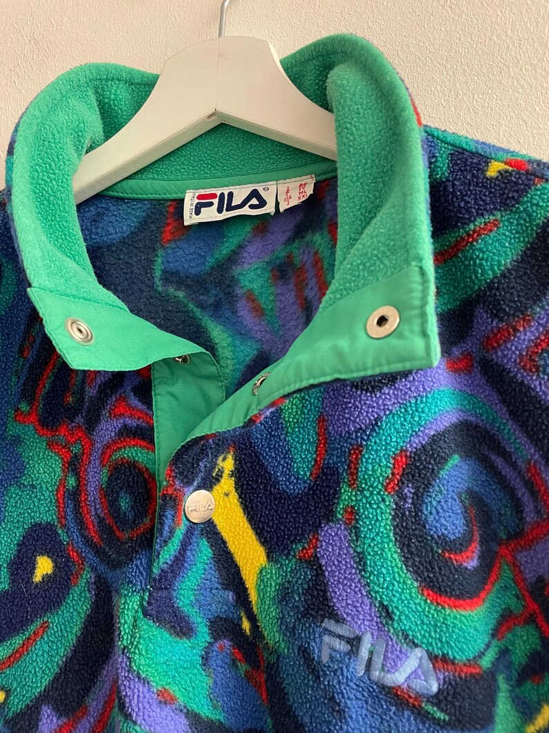 K&ouml;nnte beinhalten: Ein Fila Fleece-Pullover mit einem lebendigen, abstrakten Muster in Blau-, Gr&uuml;n-, Lila- und Gelbt&ouml;nen. Das Kleidungsst&uuml;ck verf&uuml;gt &uuml;ber einen hohen Kragen, eine kurze Knopfleiste mit silberfarbenen Druckkn&ouml;pfen und das Fila-Logo auf der Vorderseite.