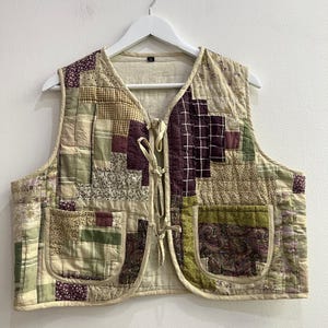 May include: A sleeveless quilted vest featuring a patchwork design of various fabrics in shades of olive green, cream, and deep plum. The vest includes two front patch pockets, a cream-colored interior lining, and three front tie closures.