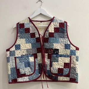 Puede incluir: Un chaleco acolchado sin mangas con un diseño de patchwork de cuadrados de tela en azul, crema y rojo oscuro con varios patrones florales y geométricos. El chaleco tiene un cuello en V, dos bolsillos de parche y finas cintas para atar al frente.