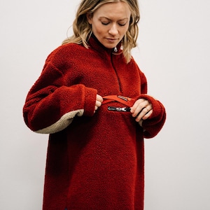 Peut inclure: Un pull en polaire rouge avec un col montant et une fermeture éclair quart de zip. Le pull a une petite poche zippée sur la poitrine et des coudières beiges. Le modèle porte le pull.