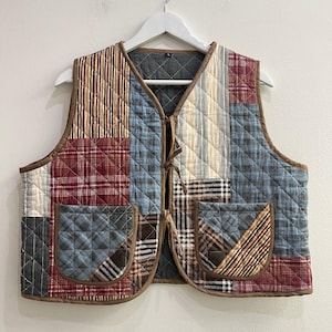 Può includere: Un gilet trapuntato con un design patchwork di vari tessuti a quadri e a righe nei toni del blu, rosso, marrone e crema. Il capo presenta uno scollo a V, due tasche applicate sul davanti, bordi marroni e due sottili chiusure a laccio.