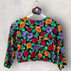 Könnte beinhalten: Ein langärmeliges Oberteil mit schwarzer Basis und einem lebendigen Blumenmuster in Rot, Orange, Lila und Grün. Das Kleidungsstück verfügt über eine einzelne Brusttasche und wird auf einem weißen Kleiderbügel mit passendem Haargummi präsentiert.