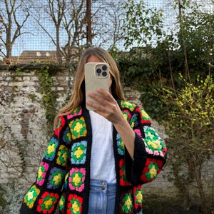 Könnte beinhalten: Eine Häkelstrickjacke mit schwarzer Basis und einem mehrfarbigen Granny-Square-Muster in leuchtenden Neonfarben wie Pink, Gelb, Grün, Blau und Rot. Das Kleidungsstück hat eine offene Vorderseite, Dreiviertelärmel und eine kastige Passform.