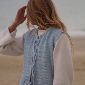 May include: A light blue knitted vest with a textured pattern and tie closures, worn over a white long-sleeved shirt. The sleeves are a solid white. The background is a beach scene with sand and ocean.