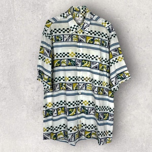 Puede incluir: Una camisa blanca de manga corta con botones y un patrón geométrico en tonos de verde, azul y amarillo. La camisa tiene cuello y cierre de botones.