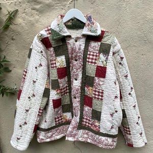 Pode incluir: Um casaco acolchoado com um design de patchwork de quadrados de tecido em creme, verde-azeitona e vermelho escuro. A peça inclui uma gola dobrada, mangas compridas com videiras florais bordadas e fecho frontal com botões metálicos.