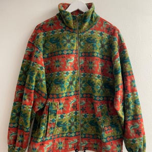 Op de afbeelding: Een fleecejack met lange mouwen en een levendig, herhalend patroon van rode, groene en blauwgroene sneeuwvlokken en abstracte motieven. Het jack heeft een ritssluiting over de gehele lengte, een hoge kraag en twee zakken aan de voorzijde.