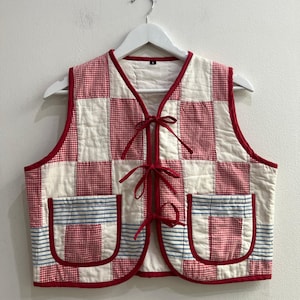 Könnte beinhalten: Eine ärmellose, gesteppte Weste mit einem Patchwork-Design aus rot-weiß karierten Vichy-Quadraten, die sich mit einfarbigem cremeweißem Stoff abwechseln. Die Weste hat rote Kanten, drei Bindebänder vorne und zwei aufgesetzte Taschen mit blau-weißen Streifen.