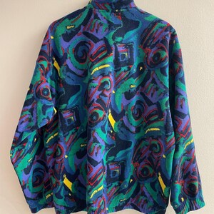 K&ouml;nnte beinhalten: Eine lang&auml;rmelige Fleecejacke mit einem lebendigen, mehrfarbigen abstrakten Muster in Dunkelblau, leuchtendem Gr&uuml;n, Violett, Rot und Gelb. Das Kleidungsst&uuml;ck verf&uuml;gt &uuml;ber einen hohen Kragen, lange &Auml;rmel mit elastischen B&uuml;ndchen und einen Rei&szlig;verschluss.