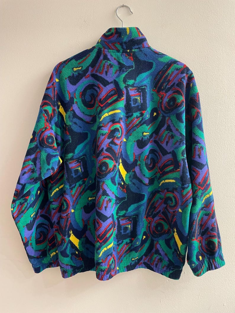 K&ouml;nnte beinhalten: Eine lang&auml;rmelige Fleecejacke mit einem lebendigen, mehrfarbigen abstrakten Muster in Dunkelblau, leuchtendem Gr&uuml;n, Violett, Rot und Gelb. Das Kleidungsst&uuml;ck verf&uuml;gt &uuml;ber einen hohen Kragen, lange &Auml;rmel mit elastischen B&uuml;ndchen und einen Rei&szlig;verschluss.