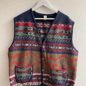 Puede incluir: Un chaleco sin mangas con botones en la parte delantera, con un patrón geométrico multicolor en tonos azul marino, rojo, verde y beige. La prenda incluye dos bolsillos delanteros de parche, un cuello en V y un borde azul marino sólido.