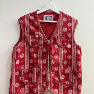 Può includere: Un gilet rosso senza maniche con una stampa bandana bianca caratterizzata da motivi geometrici a fiocco di neve e strisce verticali. Il capo presenta una chiusura centrale con zip argentata e quattro tasche applicate arrotondate con rivetti.