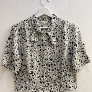Könnte beinhalten: Eine kurzärmelige Bluse mit Knopfleiste, schwarzem und weißem Blumenmuster, spitzem Kragen mit Bindedetail am Hals und hellen Knöpfen. Das Kleidungsstück wird auf einem weißen Kleiderbügel vor einem einfarbigen Hintergrund präsentiert.