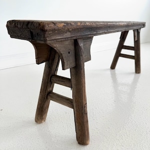 May include: A rustic, weathered wooden bench with a rectangular seat and four angled legs. The bench is made of dark brown wood, showing visible grain and signs of age. The legs are connected by horizontal supports, creating a sturdy structure.