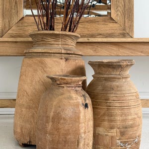 May include: Three antique wooden vases of varying sizes. The vases are a light brown color with visible wood grain and carved details. The largest vase holds dried branches. A wooden framed mirror is in the background.