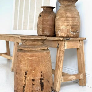 May include: Three wooden decorative vases of varying sizes and shapes sit on a rustic wooden bench. The vases are made of light brown wood with visible grain and imperfections. A large, cylindrical vase is in the foreground.