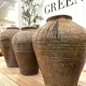 May include: Three antique brown ceramic vases with textured surfaces and a weathered appearance. The vases are arranged in a row on a wooden surface, with a blurred background featuring greenery and the word "GREEN".
