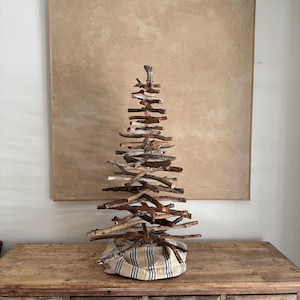 May include: A decorative Christmas tree made of natural driftwood branches. The tree is assembled on a striped fabric base and stands on a wooden dresser. The background features a neutral-toned painting.