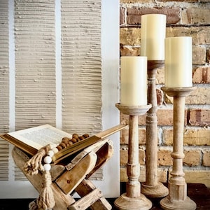 May include: A rustic wooden book stand with an open book and wooden beads, alongside three wooden candle holders with cream-colored candles. The background features a textured white wall and a brick wall.