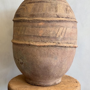 May include: A large, antique, brown earthenware vessel with a rounded base and a textured, patterned surface. The vessel is decorated with horizontal and curved lines. It sits on a small, round, wooden stool.