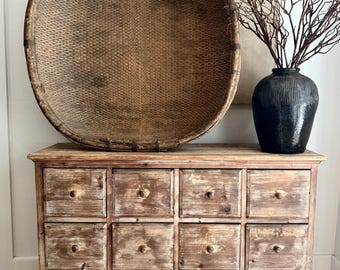 May include: A rustic wooden cabinet with twelve drawers, topped with a large woven basket and a black textured vase holding dried branches. The cabinet has wooden knobs and a distressed finish, creating a vintage aesthetic.