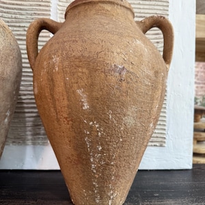 May include: A brown, antique-style vase with two handles. The vase has a textured surface and a slightly tapered shape. The rim is a darker brown color. The vase is likely made of clay or a similar material.