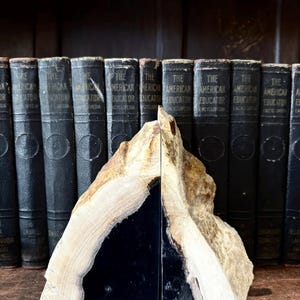 May include: A pair of natural stone bookends. One side is a polished black, the other is a mix of beige and brown tones. The bookends are in front of a row of dark books with gold lettering.