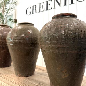May include: Three large, weathered ceramic jars with a dark, mottled olive-green glaze. Each vessel features a wide, rounded body that tapers toward a narrow base, a short neck, and a thick, circular rim. The surfaces show signs of age and texture.