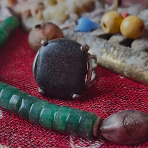 Puede incluir: Primer plano de un anillo de plata con una piedra cuadrada grande y oscura. Un collar de cuentas verdes y otras joyas están dispuestas sobre una superficie roja estampada. La piedra del anillo tiene un aspecto moteado.