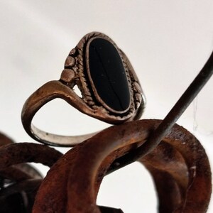 Könnte beinhalten: Ein silberner Ring mit einem schwarzen ovalen Stein, der in einer dekorativen Fassung sitzt. Der Ring liegt auf einer rostigen Metalloberfläche.