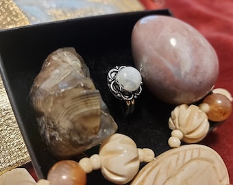 May include: A collection of natural elements including a moonstone ring, a large pink stone, a shell, and a beaded necklace. The ring has a silver band and a round, white moonstone. The pink stone is oval-shaped. The shell is brown and tan.