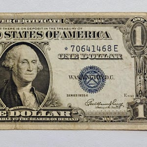 Puede incluir: Un billete de certificado de plata de un dólar de 1935, con un retrato de George Washington. El billete es de color crema con texto negro y un sello azul, y el texto dice "The United States of America."