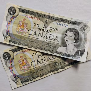 Pode incluir: Duas notas de um dólar canadense. Cada nota apresenta um retrato de uma mulher, o texto "BANK OF CANADA" e a denominação "ONE DOLLAR". As notas têm um esquema de cores creme e verde com designs e texto intrincados.