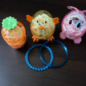 May include: Three decorative containers shaped like a chick, a bunny, and a flower pot, filled with colorful yarn. Two blue bracelets are also present. The containers are orange, yellow, and pink. The bracelets are blue.
