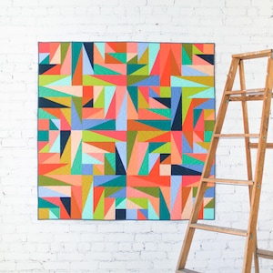 Op de afbeelding: Een kleurrijk geometrisch quilt met een modern design. Het quilt toont een patchwork van driehoeken en andere vormen in tinten rood, oranje, groen, blauw en roze. Het hangt aan een witte bakstenen muur naast een houten ladder.