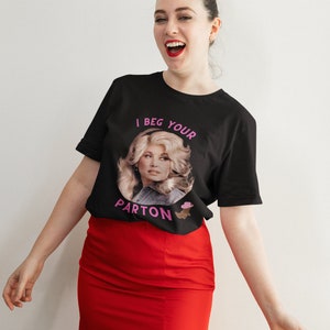 May include: Black t-shirt with a portrait of a woman and the text "I Beg Your Parton" in pink. The woman in the portrait has blonde hair and is wearing a denim jacket. The person in the image is wearing a red skirt.