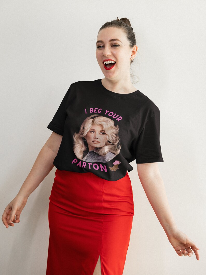 May include: Black t-shirt with a portrait of a woman and the text "I Beg Your Parton" in pink. The woman in the portrait has blonde hair and is wearing a denim jacket. The person in the image is wearing a red skirt.