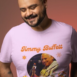 May include: Light pink t-shirt featuring the text "Jimmy Buffett" in orange retro font, with a circular image of Jimmy Buffett playing a saxophone. The shirt also has the text "Parrot Head For Life!" and flower graphics.