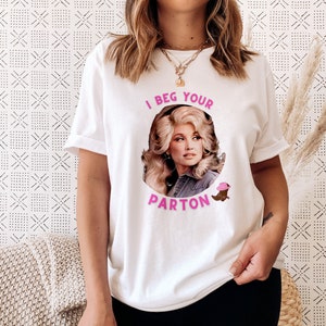 May include: White t-shirt featuring a portrait of a woman with the text "I Beg Your Parton" in pink. The design includes a pink cowboy hat and a brown boot. The shirt is a casual, unisex style.