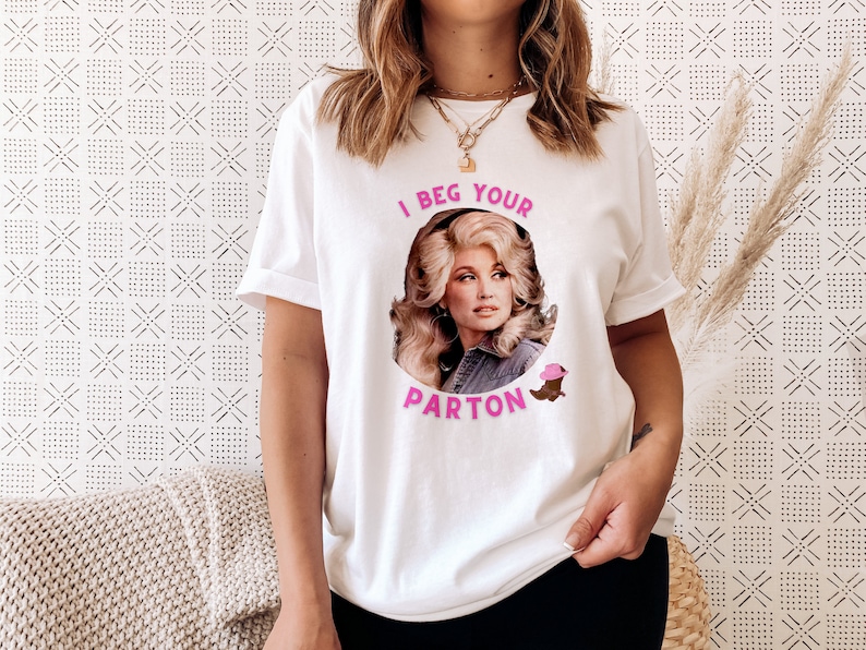May include: White t-shirt featuring a portrait of a woman with the text "I Beg Your Parton" in pink. The design includes a pink cowboy hat and a brown boot. The shirt is a casual, unisex style.