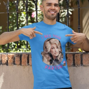 May include: A light blue t-shirt featuring a portrait of Dolly Parton with the text "I BEG YOUR PARTON" in pink. The shirt is being pointed at by a person.