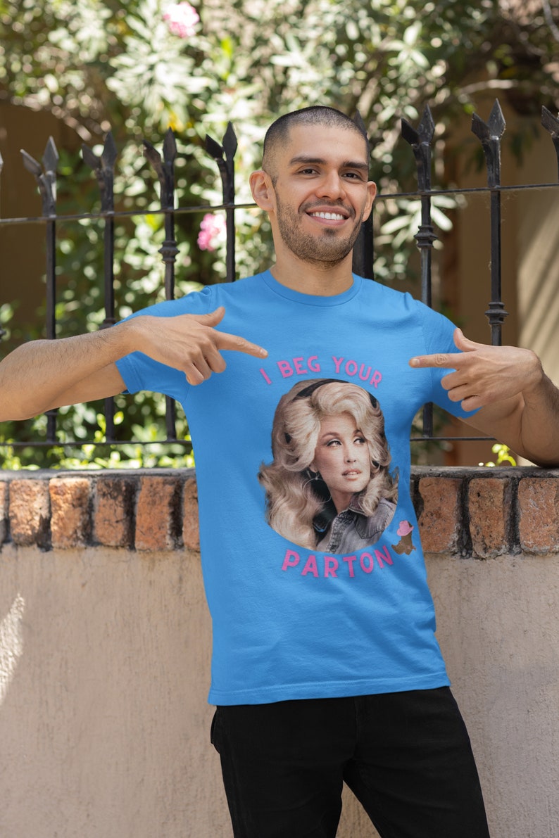 May include: A light blue t-shirt featuring a portrait of Dolly Parton with the text "I BEG YOUR PARTON" in pink. The shirt is being pointed at by a person.