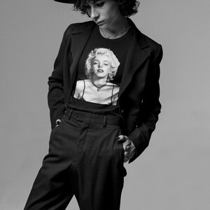 May include: Black and white image of a person wearing a black fedora hat, a black blazer, and black pants. The person is wearing a black t-shirt with a black and white image of Marilyn Monroe. The person has their hand in their pocket.