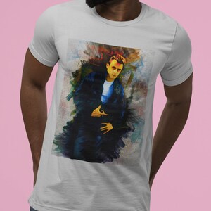 May include: A light gray t-shirt featuring a colorful artistic portrait of a person in a blue jacket and white shirt. The portrait has a painterly style with vibrant brushstrokes in shades of blue, yellow, and red.