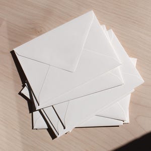 May include: A pile of white envelopes on a light wooden surface. The envelopes are rectangular with triangular flaps, creating shadows. The image is a top-down view, emphasizing the clean lines and simplicity of the stationery.