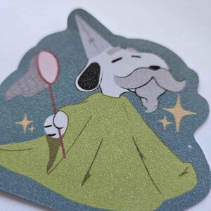 Puede incluir: Un perro de dibujos animados con una t&uacute;nica verde y un sombrero puntiagudo sostiene una red de mariposas rosa. El perro tiene una cara blanca, ojos negros, un bigote gris y una barba gris. El fondo es de color azul verdoso con estrellas dor&eacute;es.