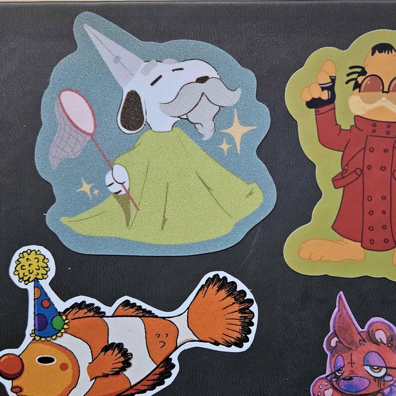 Puede incluir: Un conjunto de cuatro pegatinas de personajes de dibujos animados coloridos. Las pegatinas presentan un perro con un sombrero de mago y sosteniendo una red, un gato con un abrigo rojo y sosteniendo una pistola, un pez payaso con un sombrero de fiesta y un oso morado con una cruz roja en el pecho.