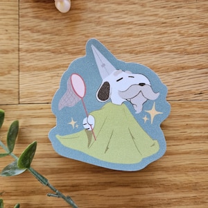 Puede incluir: Un perro blanco con una t&uacute;nica verde y un sombrero puntiagudo sostiene una red de mariposas rosa. El perro tiene barba y bigote blancos. El fondo es de color gris azulado con estrellas doradas brillantes.