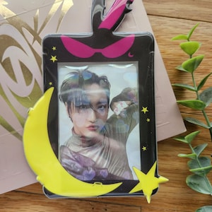 May include: A black and yellow photo card holder with a crescent moon and star design. The holder has a clear plastic window to display a photo. The photo shows a person with dark hair and a light-colored shirt.