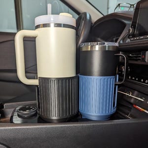 May include: Two insulated travel mugs in a car cup holder. One is cream-colored with a black base and handle, the other is black with a blue base and handle. Both have lids and are designed to keep beverages hot or cold.